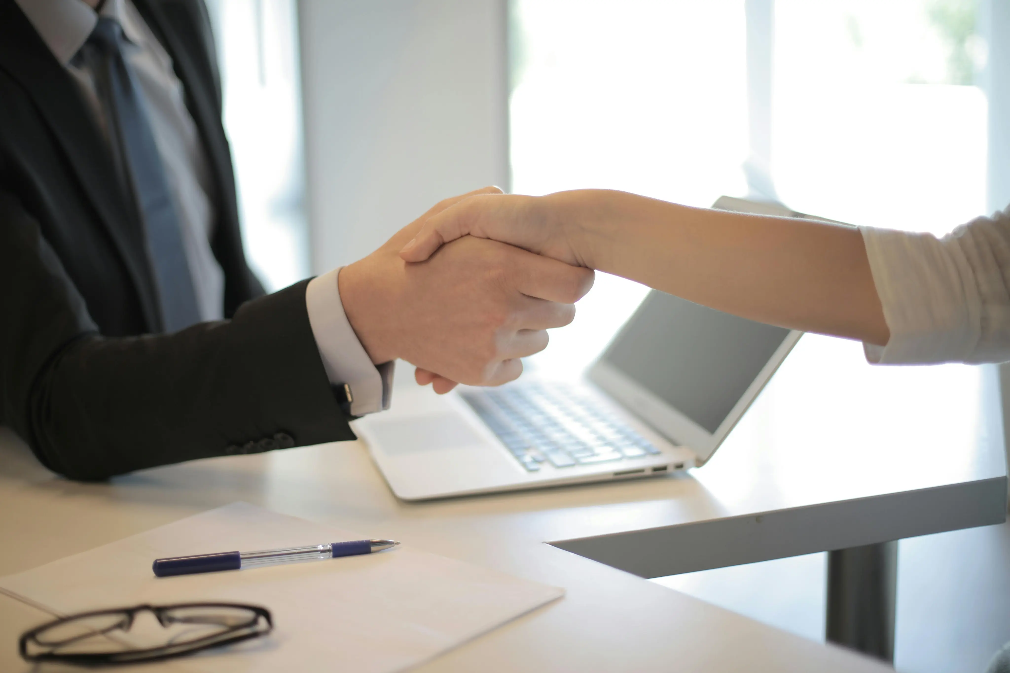 Two business professionals shaking hands over a desk with a laptop.